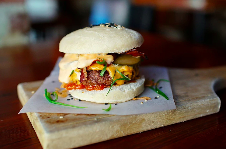 A burger served on a steamed bun and topped with a kimchi cheese and bacon