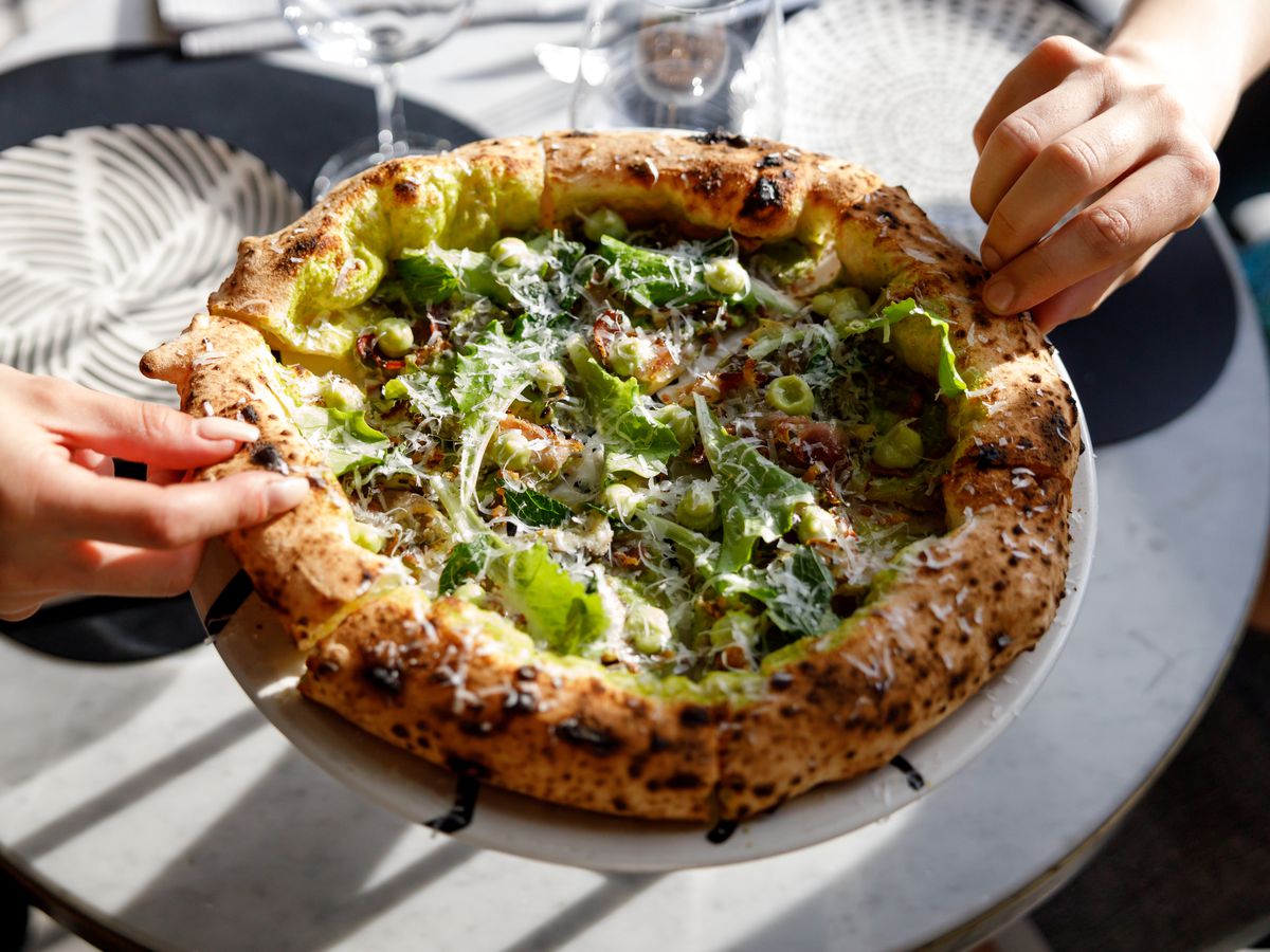 Two people pull slices from a large pizza topped with greens and cheese.