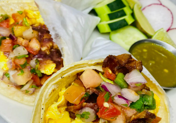 A close-up photo of two breakfast tacos stuffed with eggs, meat, and vegetables, plus sides of cucumber, lime wedges, and a green sauce.