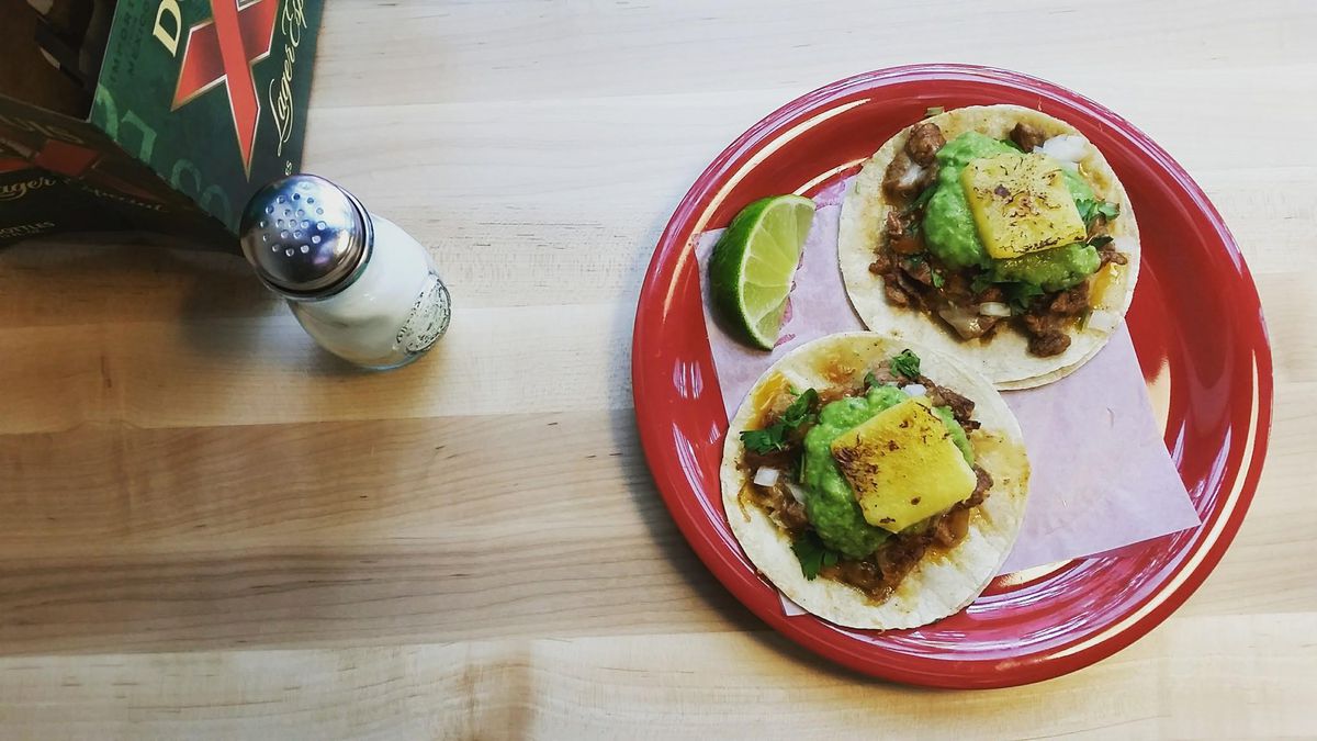 A bird’s eye view of adobada pork tacos with guacamole and slices of pineapple on a wooden table, alongside a salt shake.