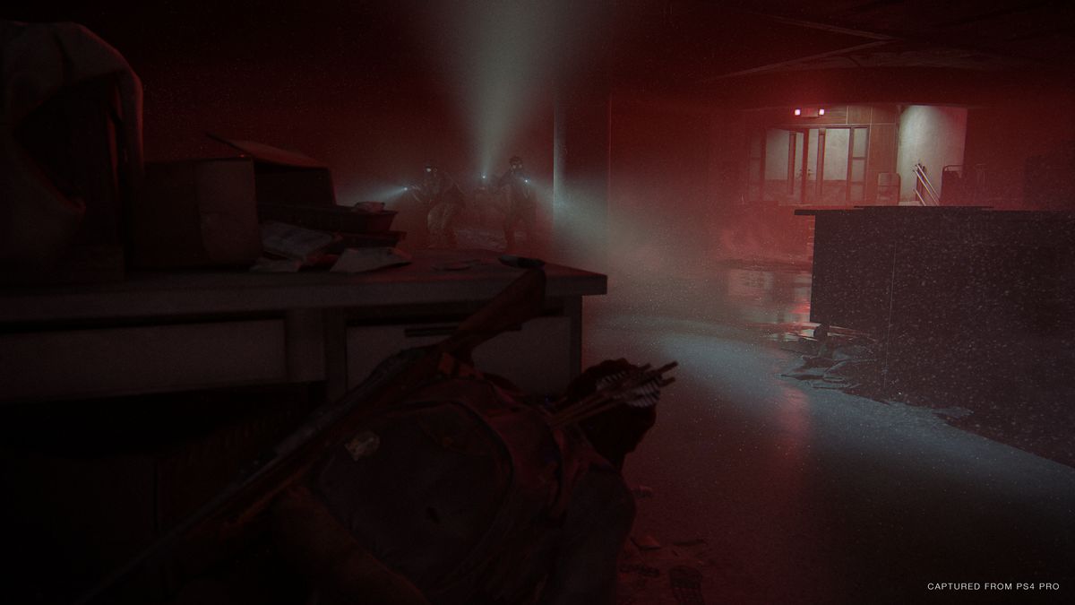 Ellie crouches behind a desk, hiding from a group of enemies in a room lit only by their flashlights and the red glow of an exit sign in The Last of Us Part 2