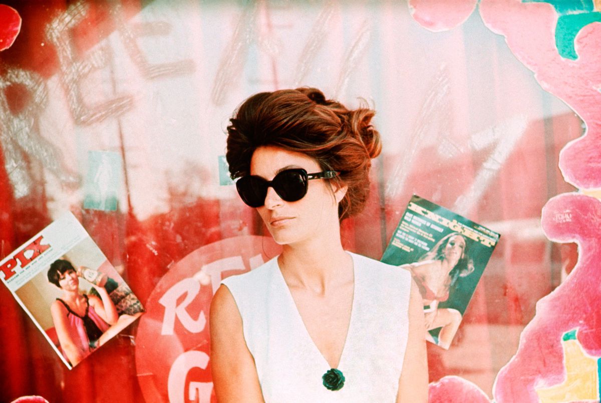 Anouk Aimée in sunglasses standing in front of a glass window with magazines plastered on the surface in Model Shop.