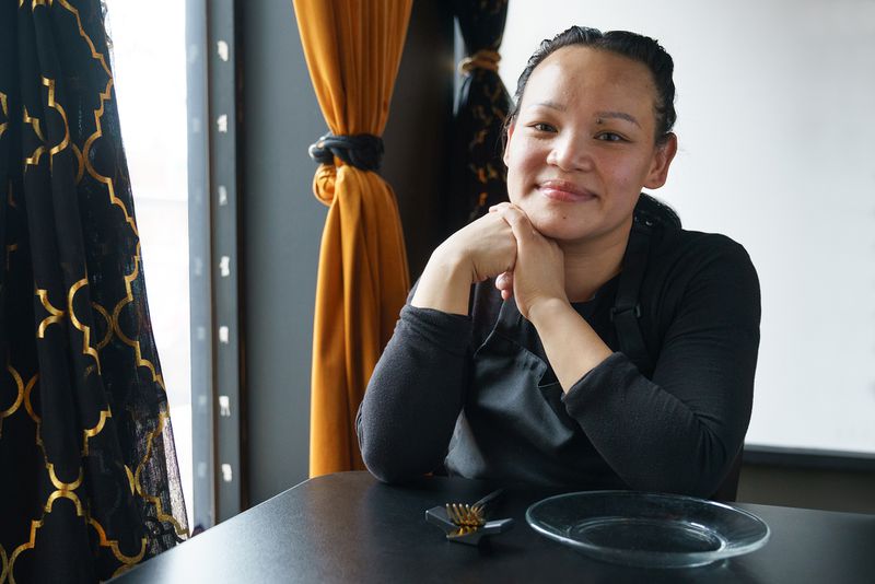 A smiling Cambodian woman wearing a black shirt and apron has her hands folded at the side of her face.