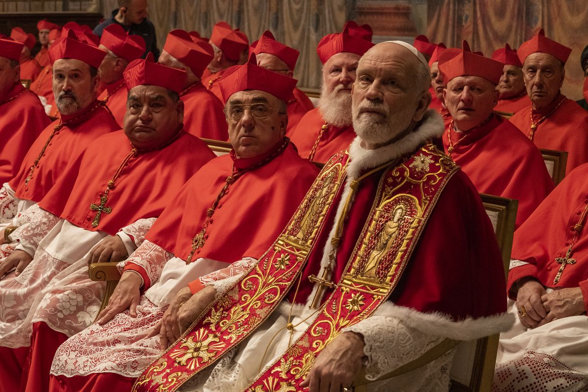John Malkovich sits wearing a white skullcap, red-and-white robes, and an elaborately gold-embroidered stole. He’s surrounded by cardinals in peaked crimson caps and red-and-white robes.
