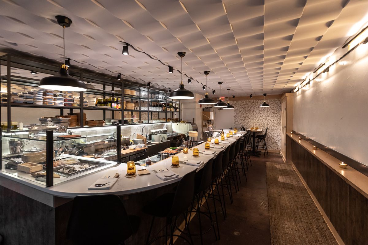 A wide-angle shot of a long curved white dining counter with table settings and a glass window surrounding a raw bar. The ceiling is covered with white wave panels and black pendant lights. 