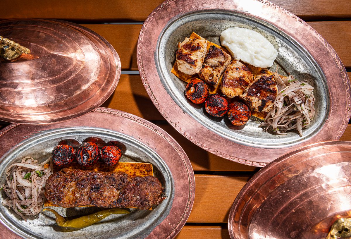 An overhead photograph of kebabs beside grilled onions and tomatoes on worn metal serving dishes.