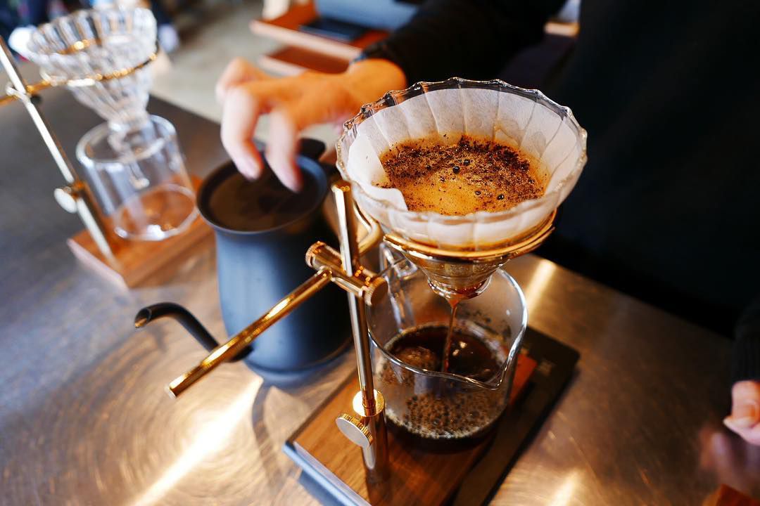 A pour over contraption with a glass beaker of coffee below and a barista blurred in the background