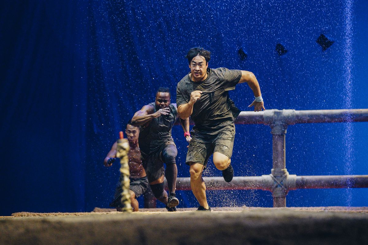 Contestants running dramatically in a still from Physical 100. Water is being sprayed down on them
