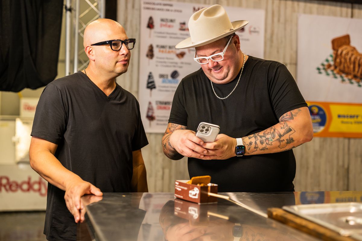 Graham Elliot takes a photo of a dessert on a stick with his iPhone. To the left, a man looks forward, ignoring the scene. 