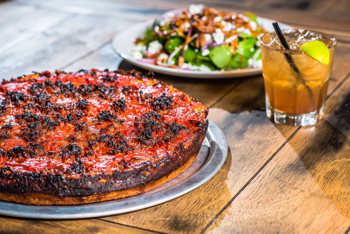 A view of a meaty Chicago deep dish-style pizza, with a cocktail and salad in the background.