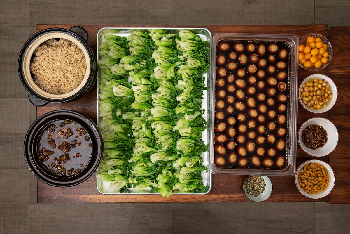 Sheet pans filled with bok choy and cooked eggs sit alongside bowls of rice, stewed meats, and other accompaniments on a tabletop.