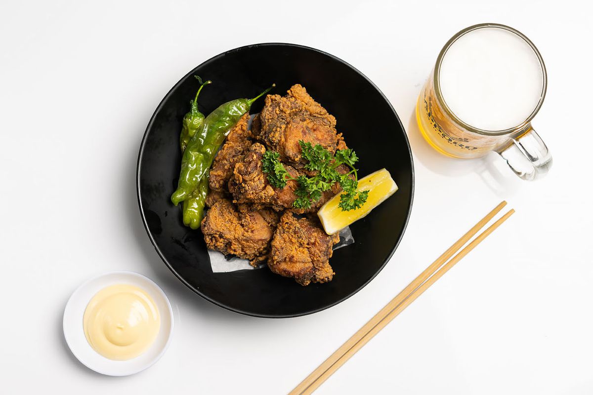 A black plate holds pieces of fried chicken as well as garnishes including lemon, parsley, and peppers. A beer, chopsticks, and creamy dipping sauce sit nearby.