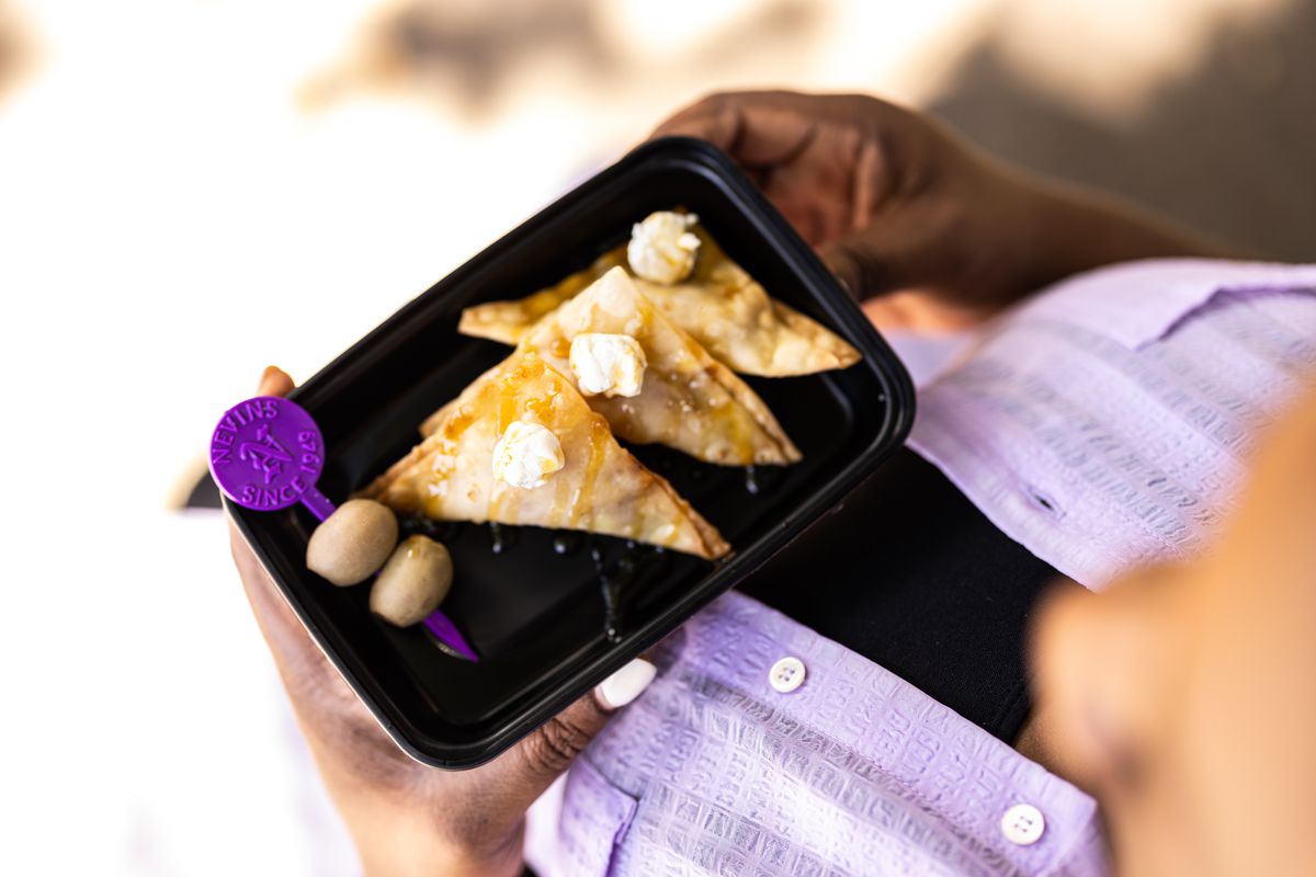 A camera looks down on a dish held by a woman, in a black plastic to-go container. It holds three wontons and a pair of olives that are speared.