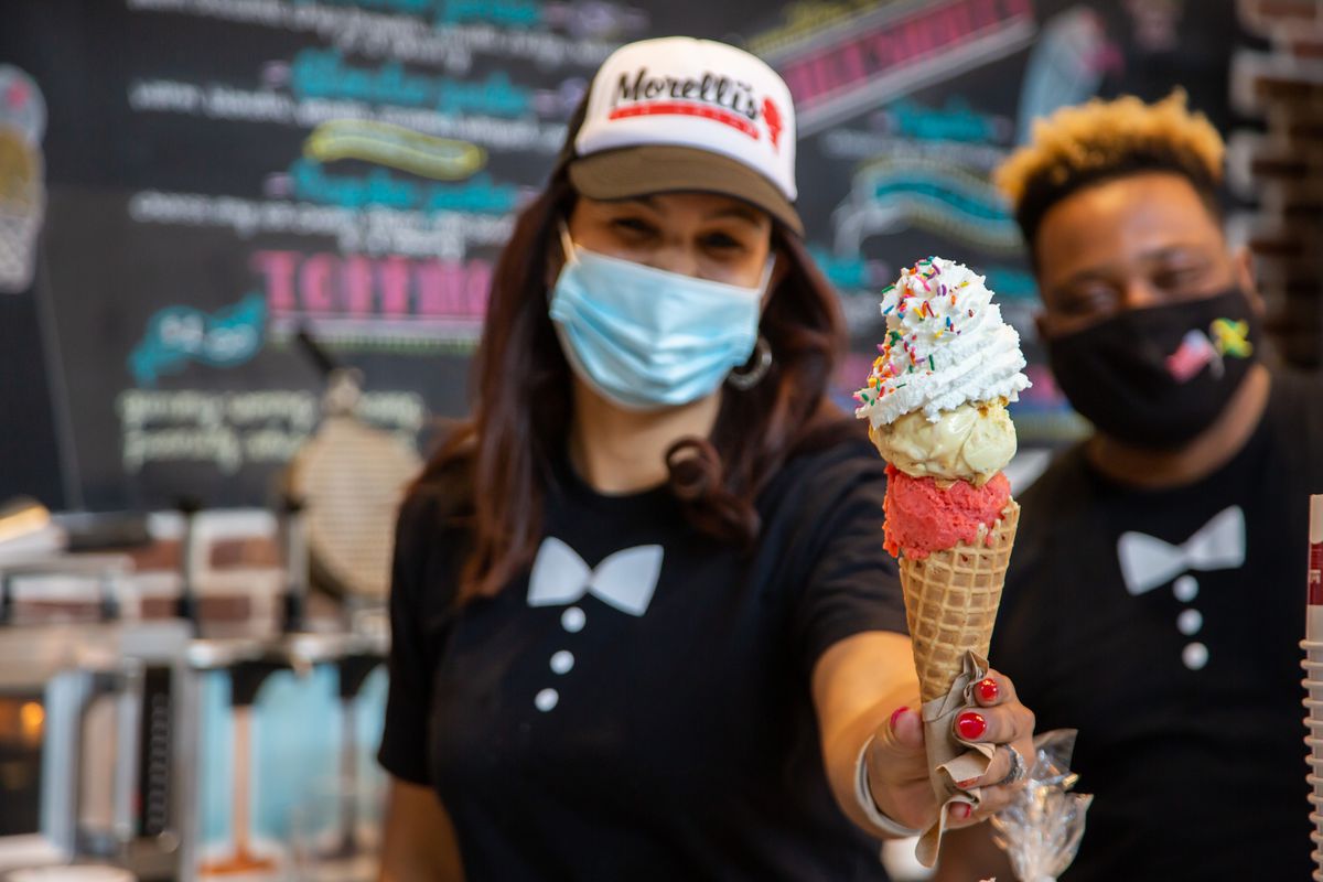 A woman with long brown hair wearing a black bow tie tee and a black and white trucker cap with the Morelli’s Ice cream logo on the front smiles behind her blue mask while holding a double scoop of strawberry and vanilla ice cream cone topped with whipped cream and rainbow sprinkles. Her coworker stands behind her in a black mask with an American flag and a Jamaican flag on it and smiles from behind their mask wearing the same bow tie tee