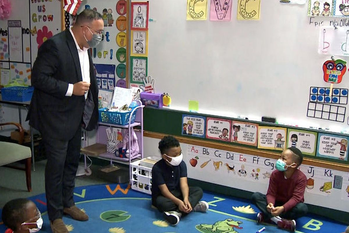 Secretary of Education Miguel Cardona, wearing a mask and standing, speaks in a classroom with elementary students, who are sitting on a rug and looking up at him.