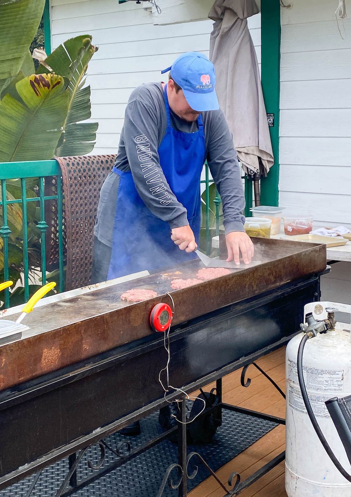 A man in blue hat and apron presses on a beef patty on a griddle.