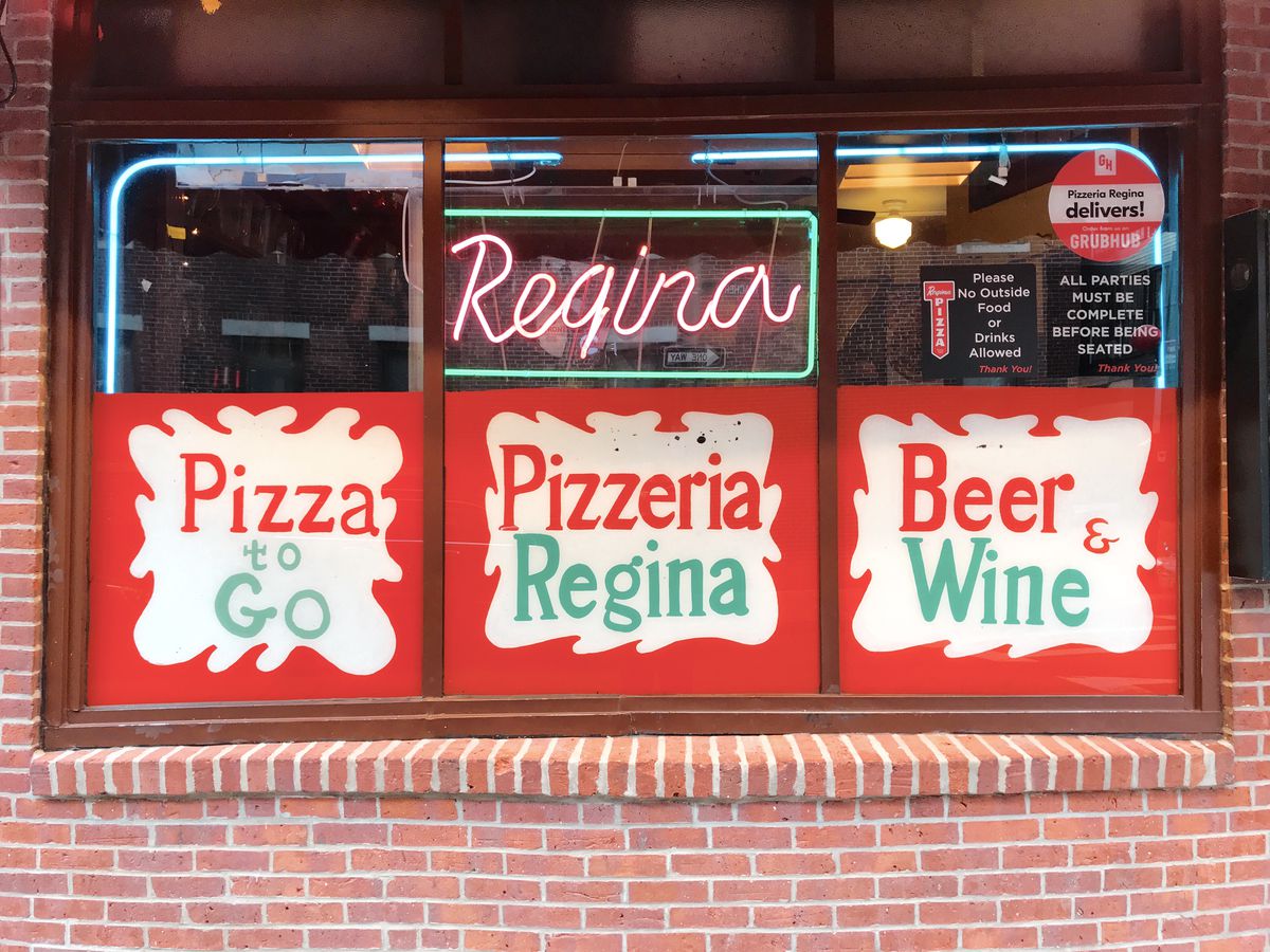 A close shot of the exterior of Pizzeria Regina’s original North End location, feature red, white, and green signs that read “Pizza to Go,” “Pizzeria Regina,” and “Beer &amp; Wine.” There’s also a red and green neon sign that says “Regina.”