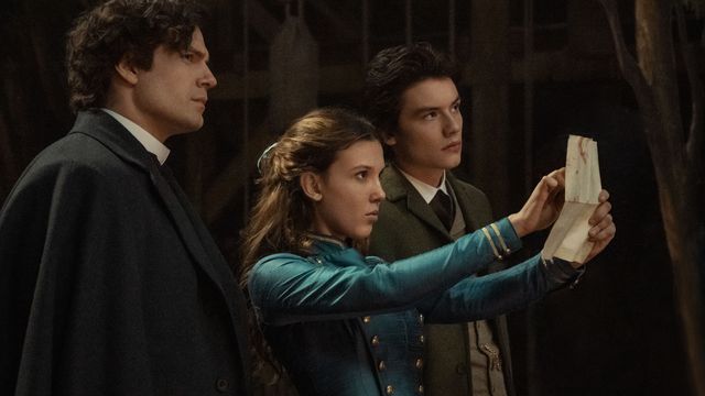 Henry Cavill as Sherlock Holmes, Millie Bobby Brown as Enola Holmes, Louis Partridge as Tewkesbury in Enola Holmes 2. Enola holds up a piece of paper and points at it while the others look on.