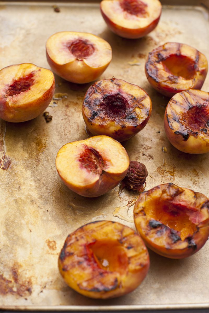 Several grilled peaches sit cut-side up on a baking tray, with grill marks visible on some of the peaches.