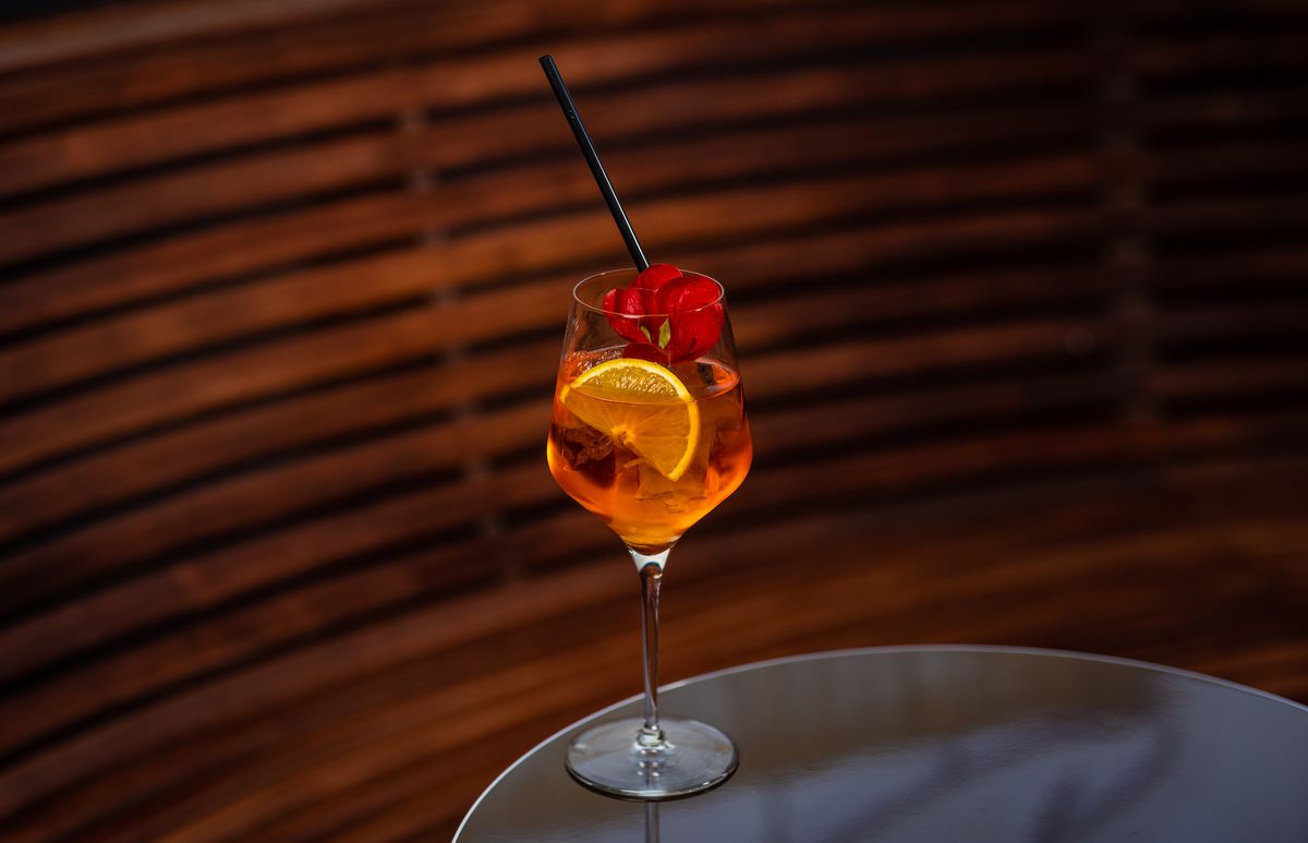 A light orange cocktail in a wine glass with floating rose petal against a wood backdrop at a new bar.
