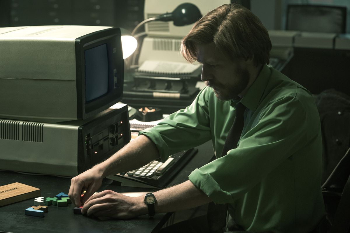 A red-haired young man with a beard plays with tetromino puzzle pieces on his desk next to an old-timey computer