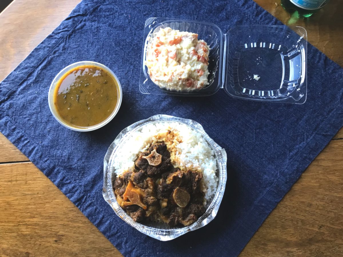 A round foil container with white rice and stewed oxtail with side of beans and sweet potatoes on a blue table cloth over a wooden table.