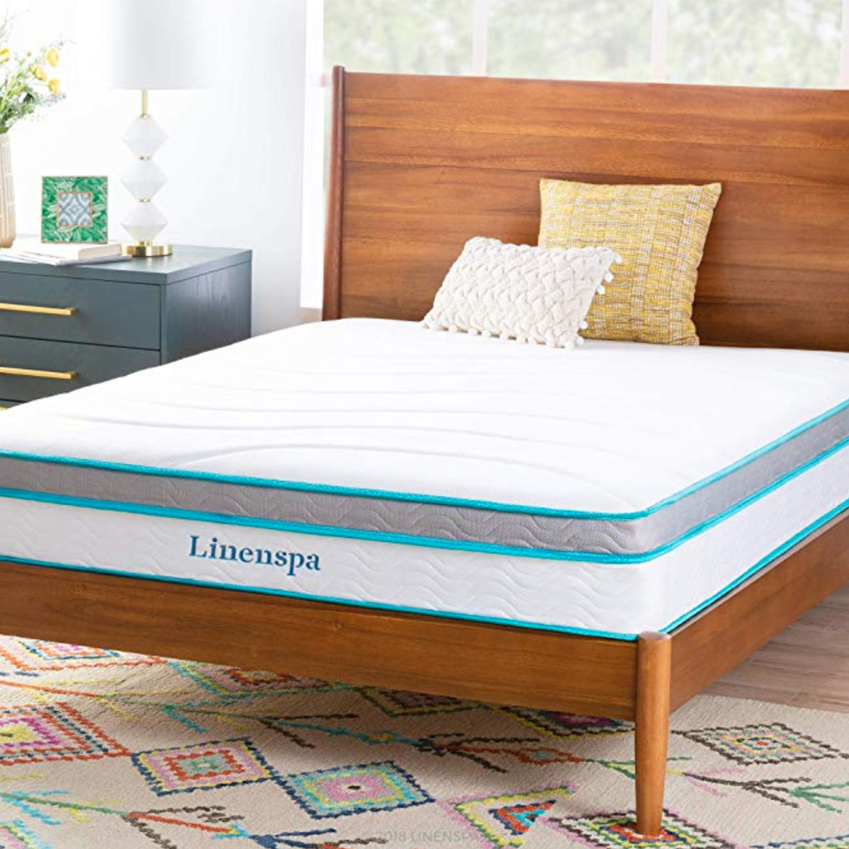 A white uncovered mattress sits on a wooden bed platform and headboard. A colorful patterned rug is underneath. 