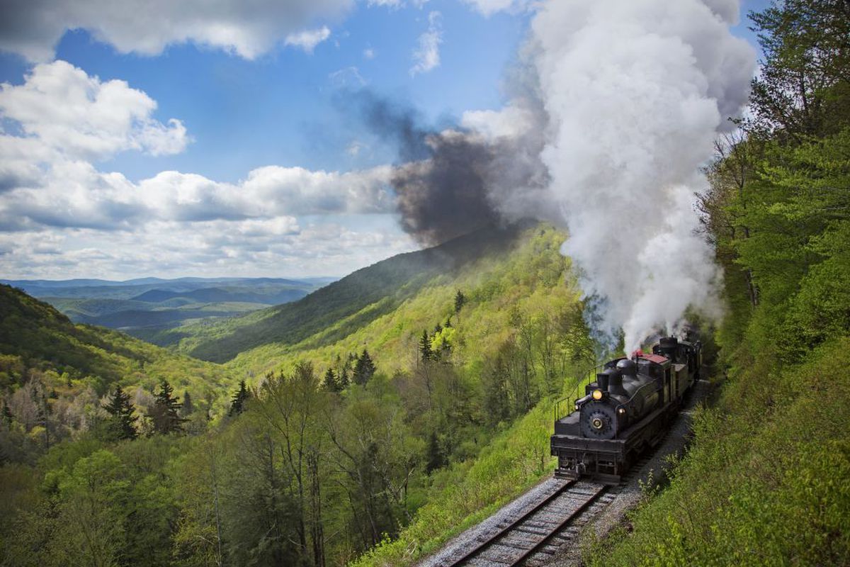 A black steam engine train spewing smoke winds through lush forests with mountain ranges in the background.