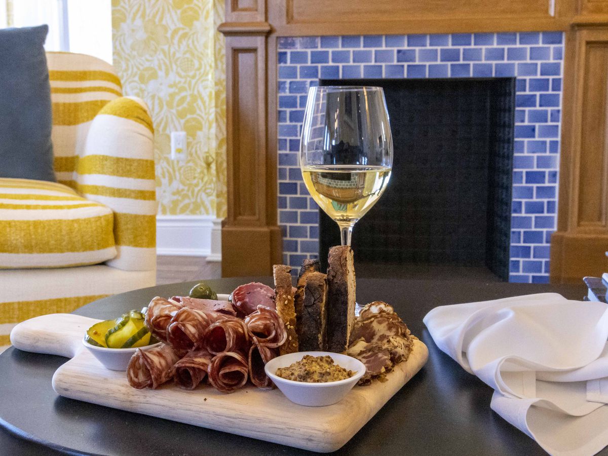 Charcuterie on a wooden cutting board next to a glass of white wine with a blue-tiled fireplace and white and yellow seating in the background