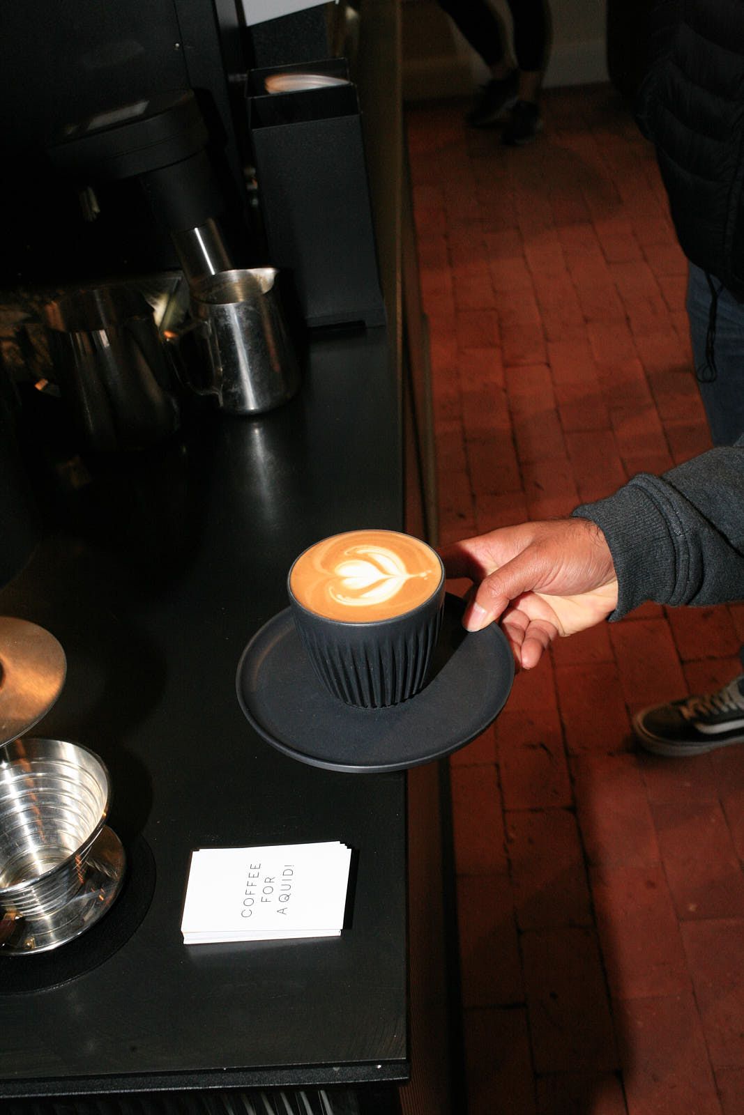 A hand picks up a flat white coffee in a black cup, the drink adorned with a latte art heart.