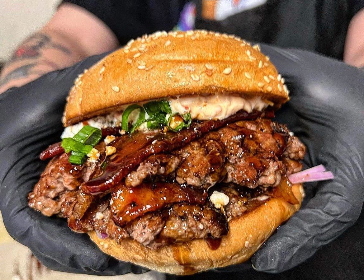 Two hands wearing black gloves holding a burger with multiple patties, goat cheese, candied bacon, sun-dried tomato, and red and green onion, with balsamic glaze.