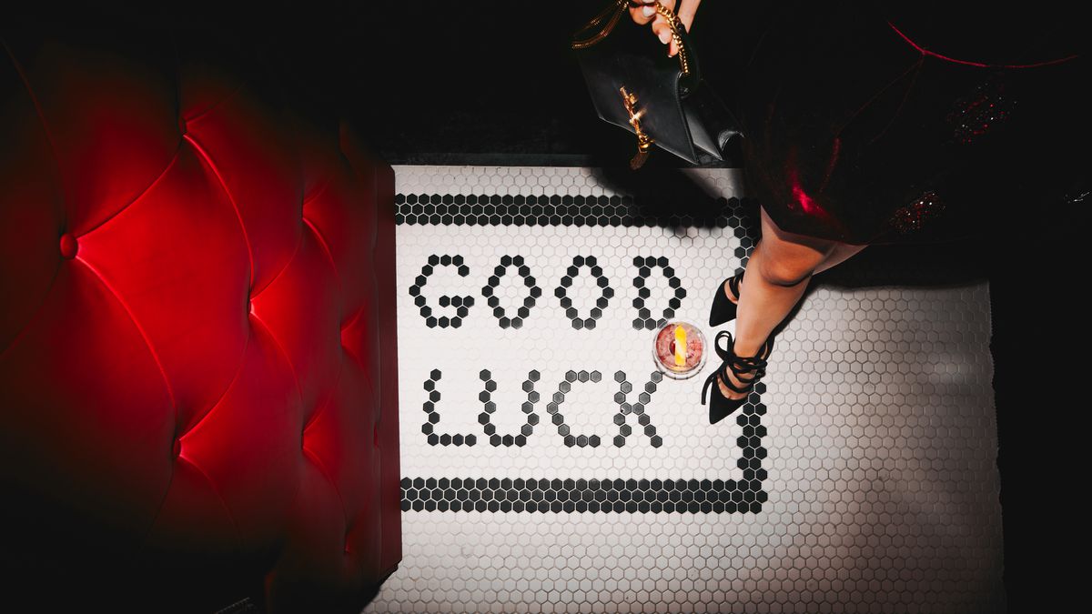A woman’s foot steps on a white tile floor. In letters made of black tile are the words “good luck.” To the left of the sign and her feet is a door covered in tufted red velvet.
