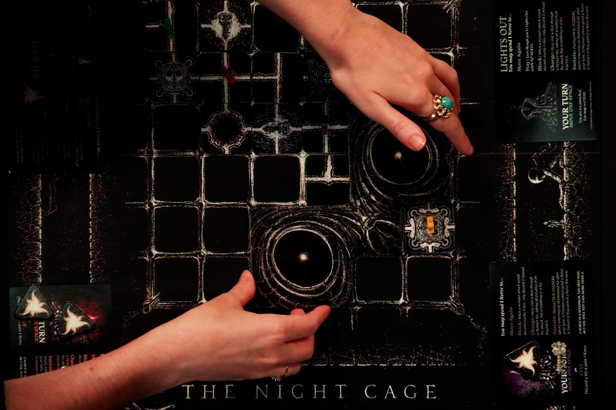 A pair of hands places a cardboard game piece for the game The Night Cage on top of the game board, which looks like a black and white 6 x 6 grid with a number of tiles and game pieces on it.