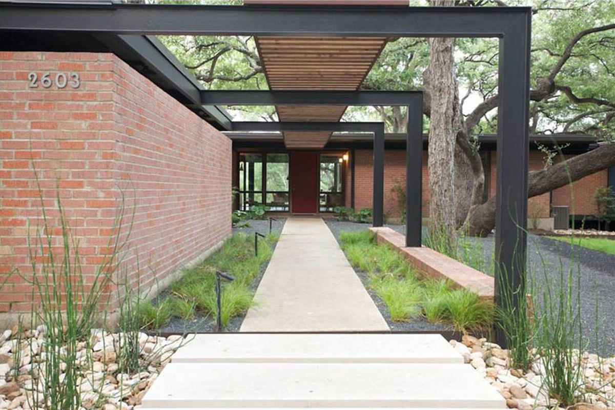 Exterior entrance to a midcentury/international style house, long walkway with wooden covering mirroring sidewalk above, red brick one-story, flat-roofed house to left and continued in back, front door and windows that enable view through house, tree over