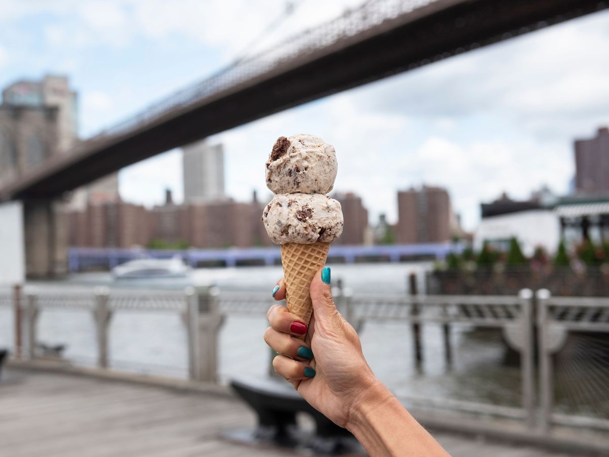 A hand holds an ice cream cone with two scoops near the waterfront in NYC.