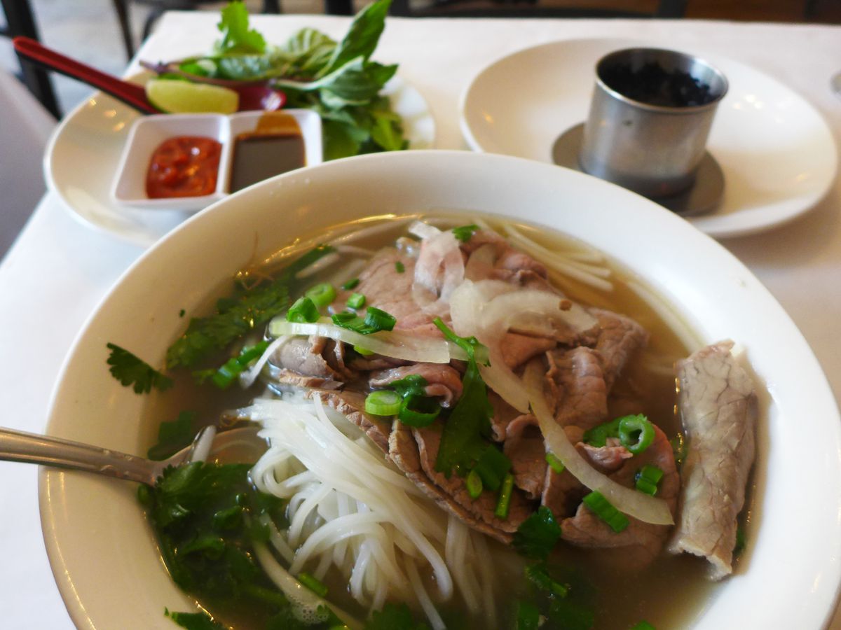A bowl of pho front and center, with a spoonful of rice noodles raised up, and a plate of basil and cilantro in the background.