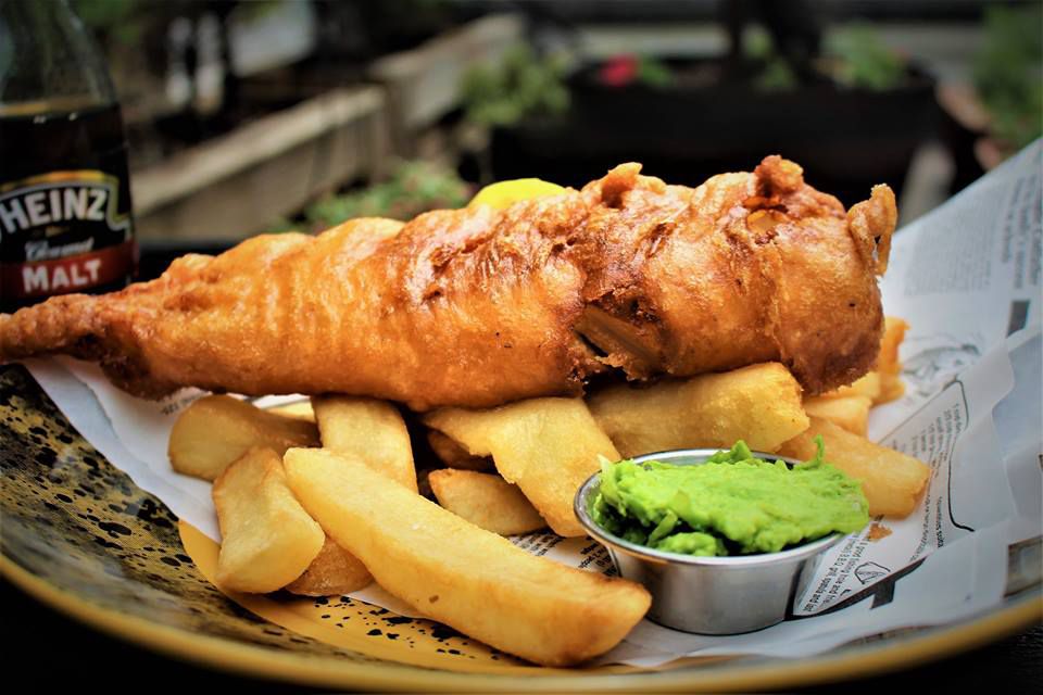 A fried piece of fish on top of thick chips with a little container of mushy peas.