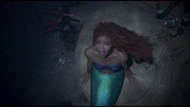 Halle Bailey as Ariel sitting on a rock and looking up while she sings