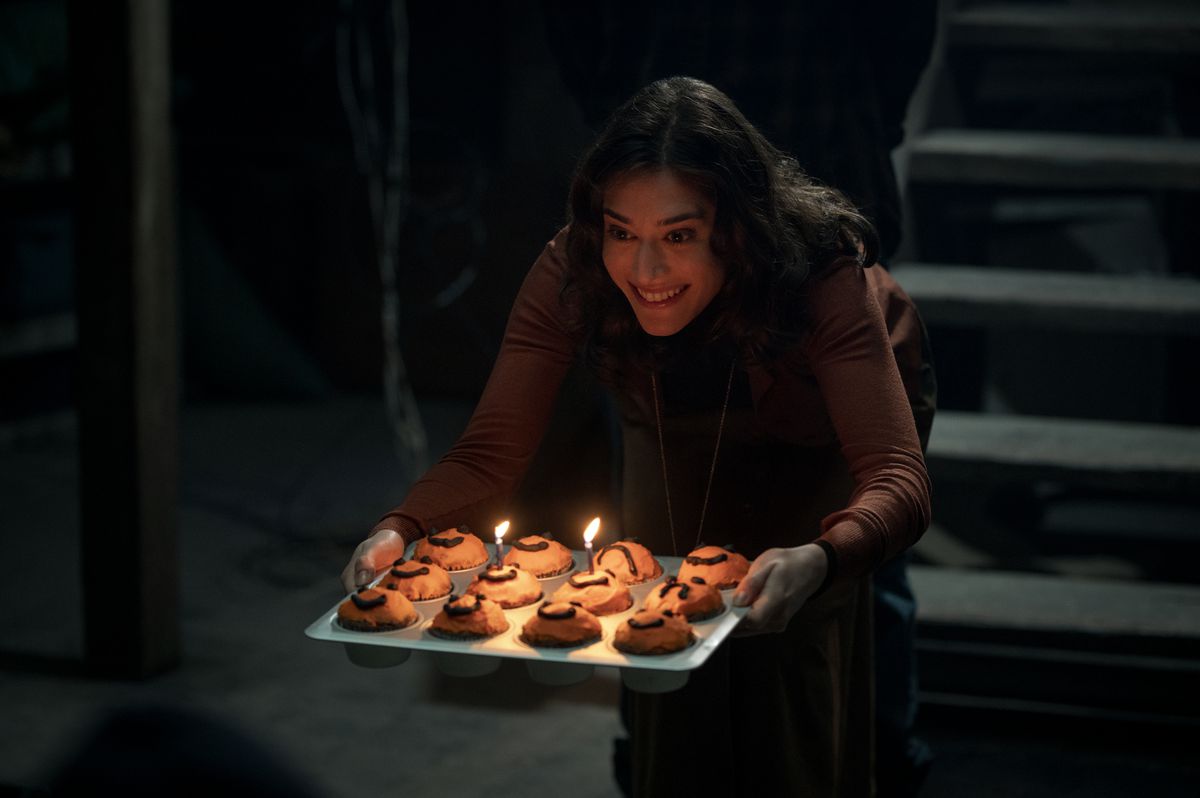 Lizzie Kaplan holds a muffin tin with candles on it while smiling creepily at the spider web.
