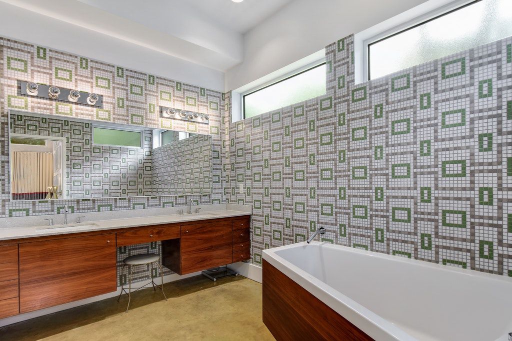 A bathroom with a green and gray geometric tile on the walls, white and wood tub.