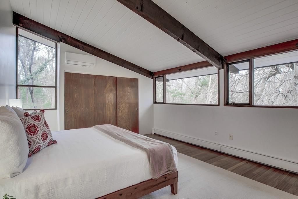 A small bedroom with a bed beneath a sloping ceiling and thin windows up top overlooking woodlands. 