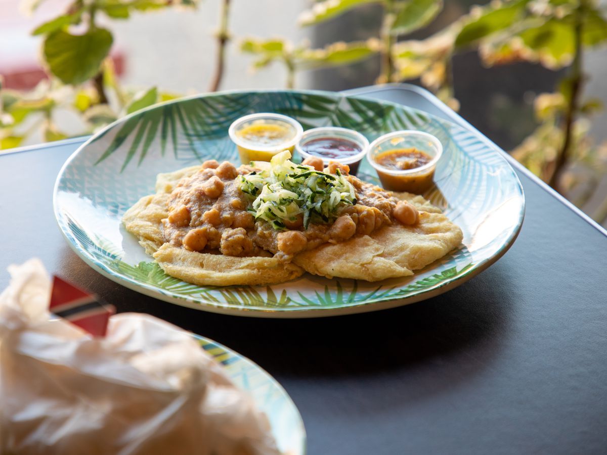 A green and blue plate holds two pieces of fried flatbread topped with a chickpea filling and cucumber salad next to three sauces on a table.