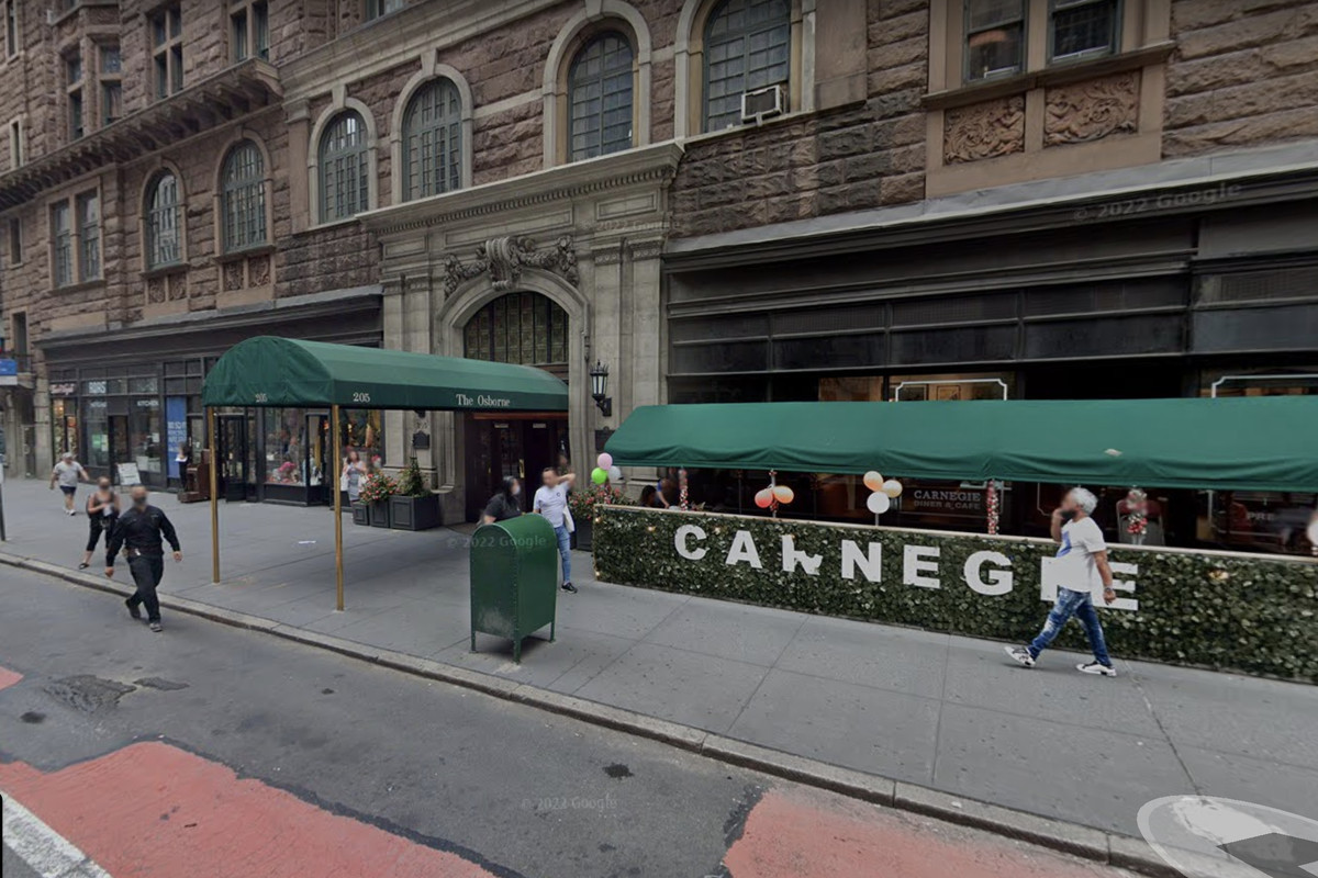 People walk by an apartment building with a green awning out front and outdoor restaurant seating to the right.