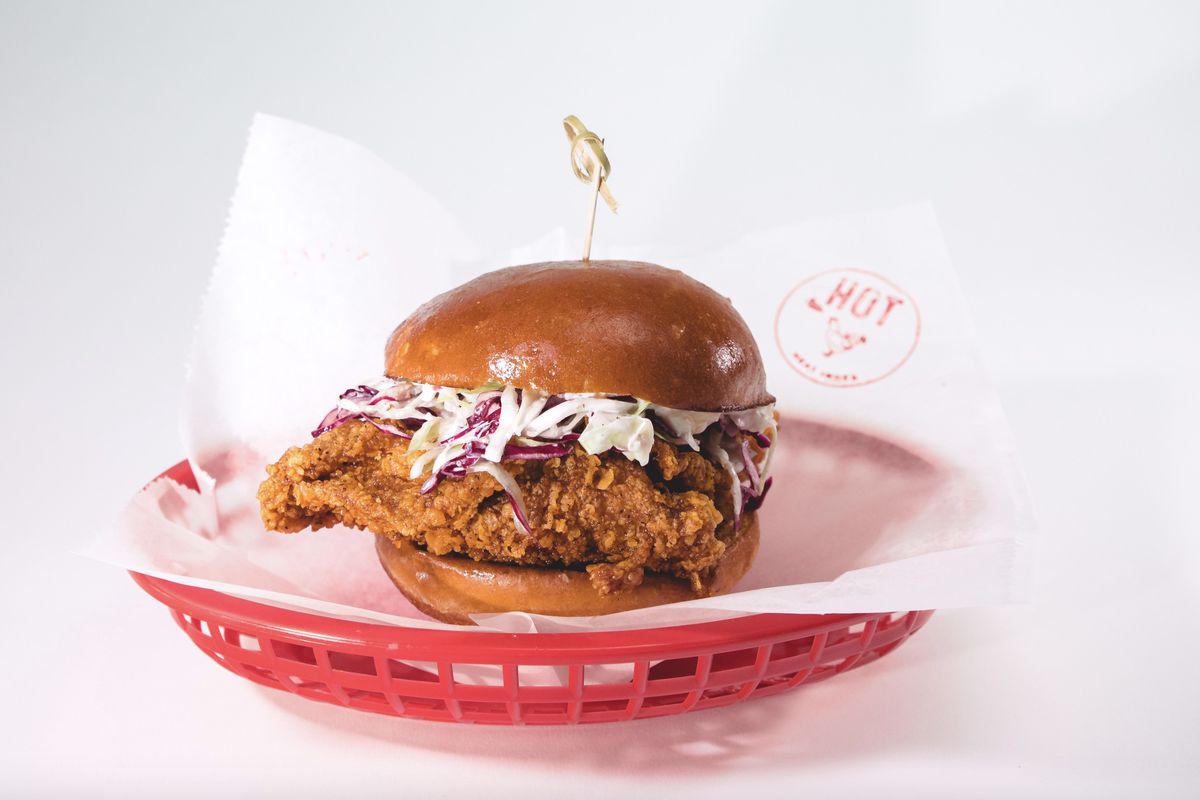A fried chicken sandwich topped with cole slaw on a brioche bun and secured with a toothpick. The sandwich sits inside a red plastic basket lined with paper. 