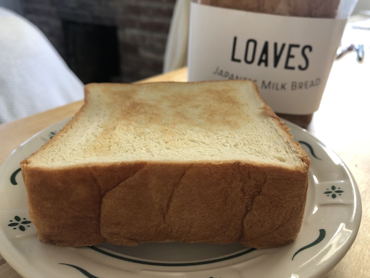 A thick slice of toasted bread sits on a small plate with a loaf of bread in the background.