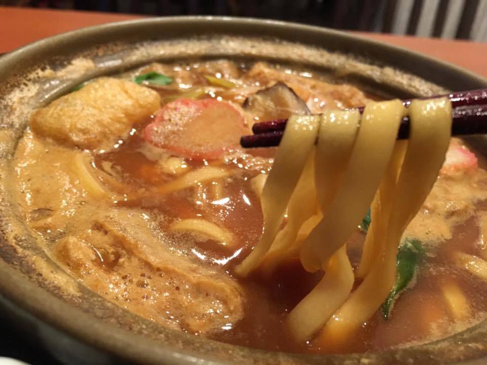 Chopsticks lift noodles from clay pot full of broth, meat, tofu, and vegetables