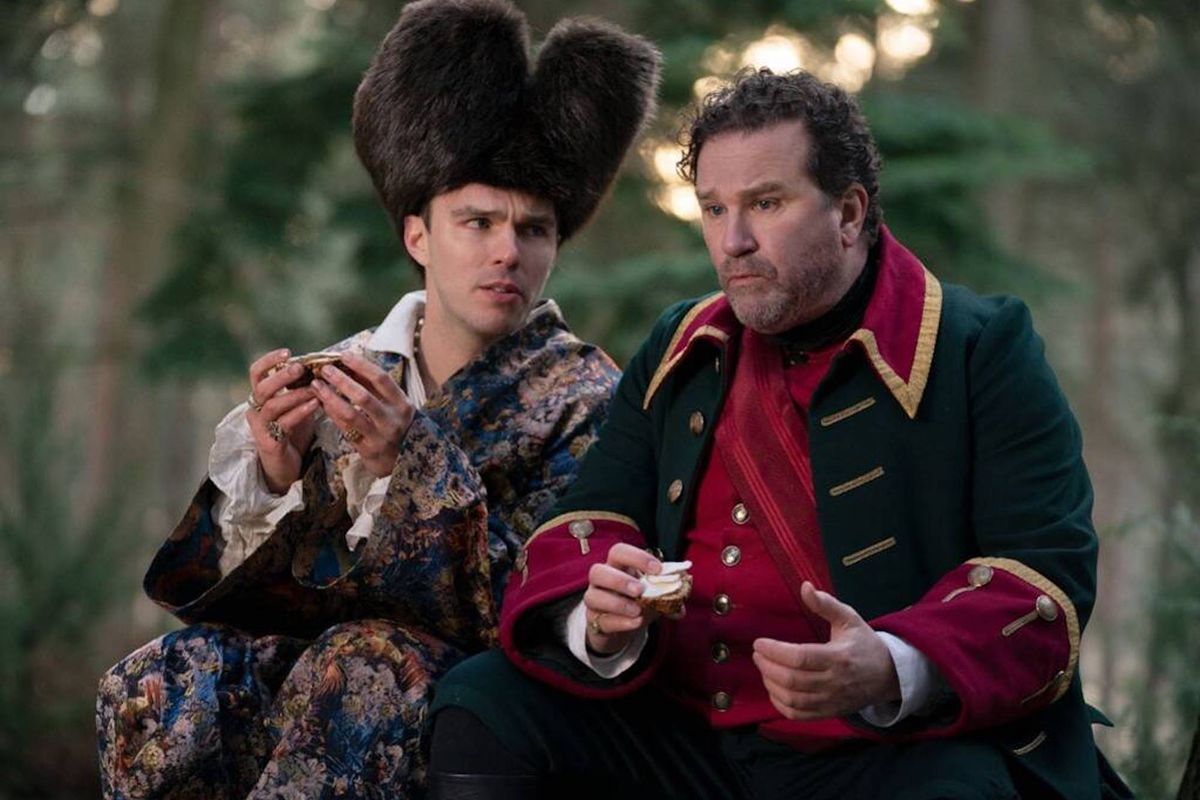 The characters Peter (played by Nicholas Hoult), dressed in a tall fur hat and brocaded coat, and General Velementov (Douglas Hodge), in an 18th century military uniform, sit in the woods eating cheese on bread.