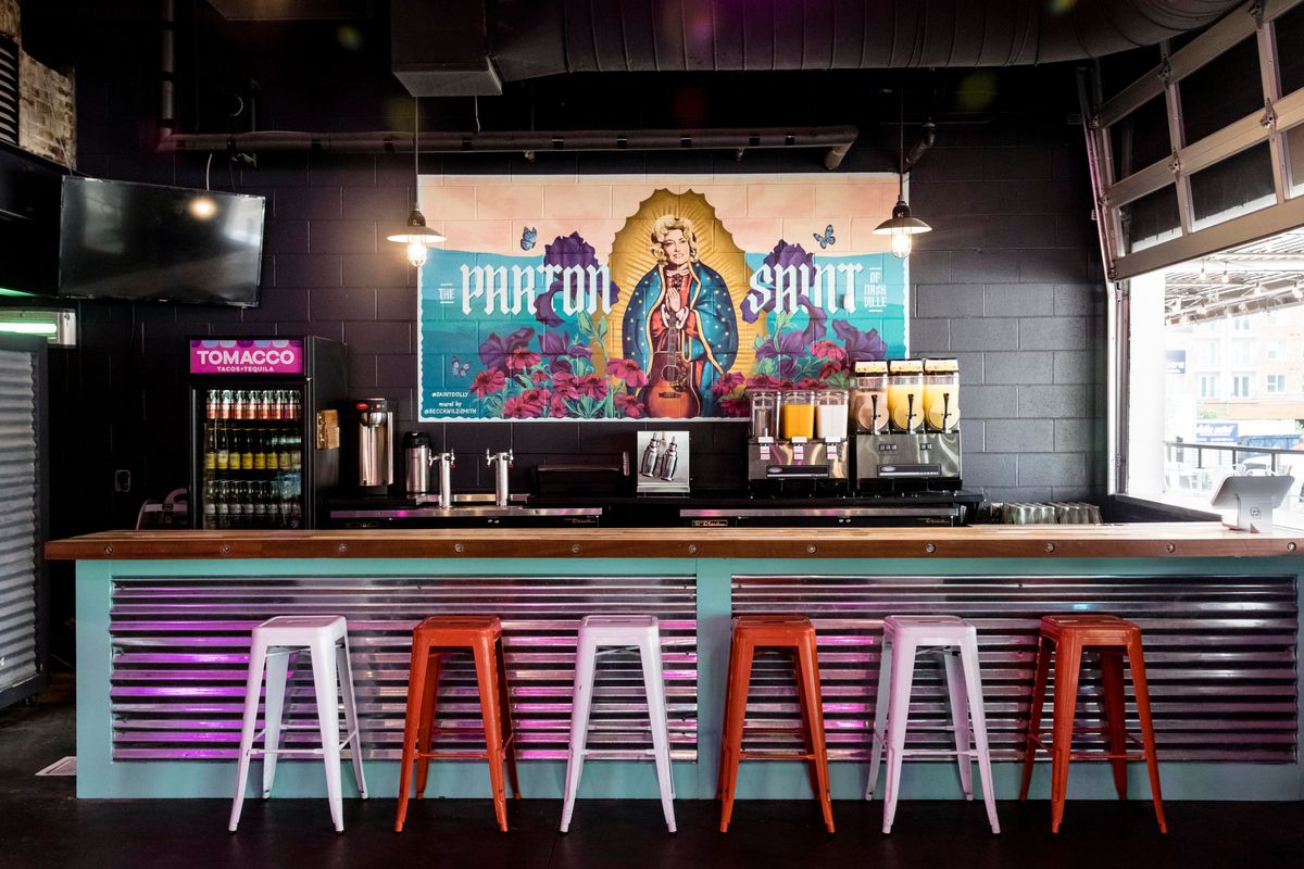 multi-colored bar stools in front of a purple metal covered bar, in front of dolly parton mural and drinks