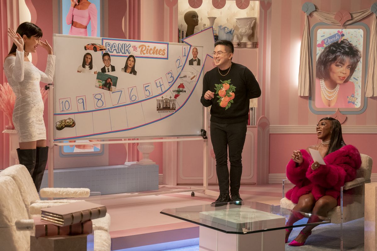 Bowen Yang and Patti Harrison play “Rank the Riches” on Ziwe, with Ziwe sitting in a chair in a fluffy red coat. Yang and Harrison stand in front of a white board that has a chart from 10 to 1 and images of rich people on it. A framed picture of Oprah Winfrey hangs in the background of the pink set.