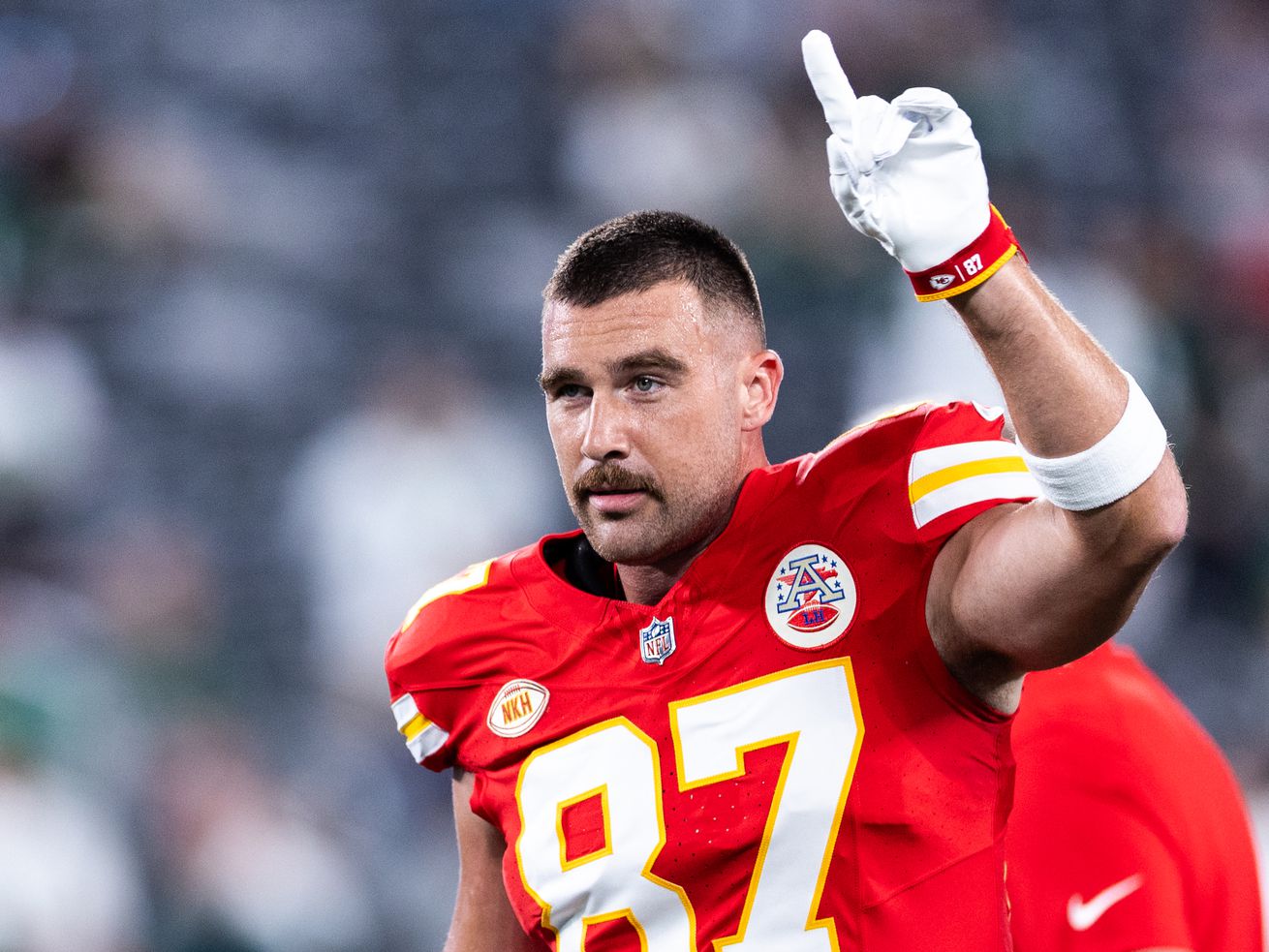 Travis Kielce, wearing a Kansas City Chiefs uniform number 87, walks off the field raising one white-gloved hand pointing to the sky.